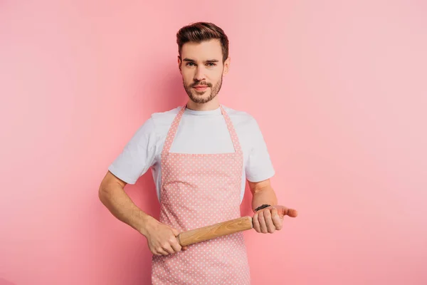 Confident young man in apron holding wooden rolling pin on pink background — Stock Photo