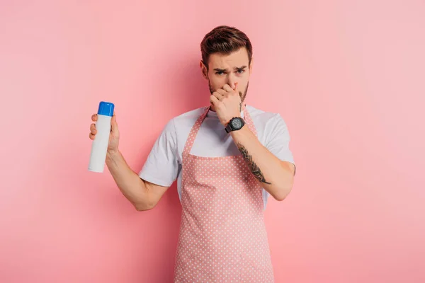 Scontento giovane uomo in grembiule tappo naso con mano mentre tiene deodorante per ambienti su sfondo rosa — Foto stock