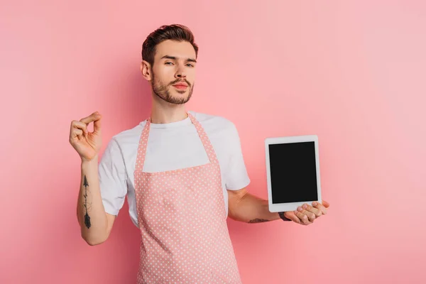 Zufriedener junger Mann in Schürze zeigt digitales Tablet mit leerem Bildschirm auf rosa Hintergrund — Stockfoto