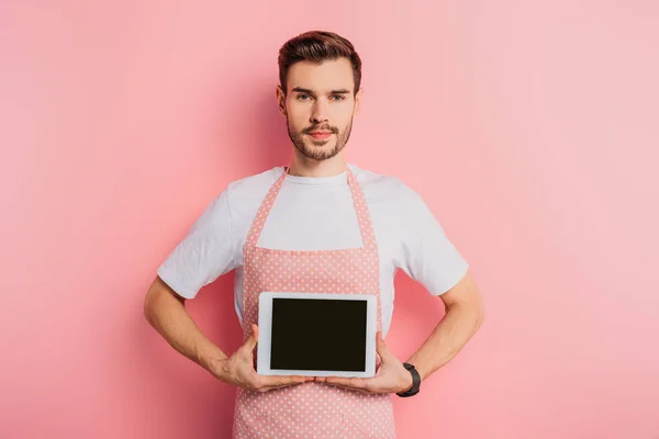 Ernster junger Mann in Schürze zeigt digitales Tablet mit leerem Bildschirm auf rosa Hintergrund — Stockfoto