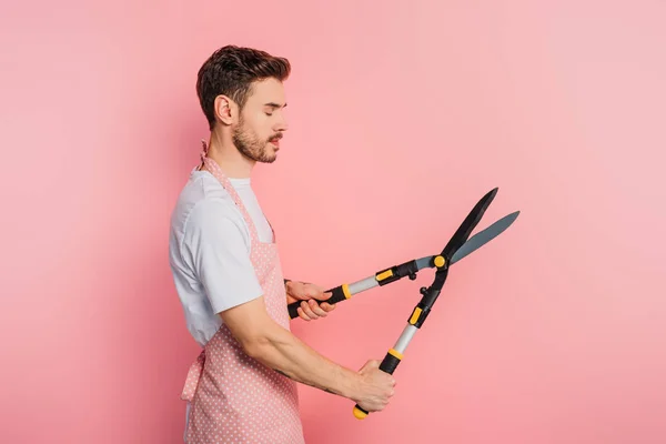 Side view of young man in apron holding gardening scissors with closed eyes on pink background — Stock Photo