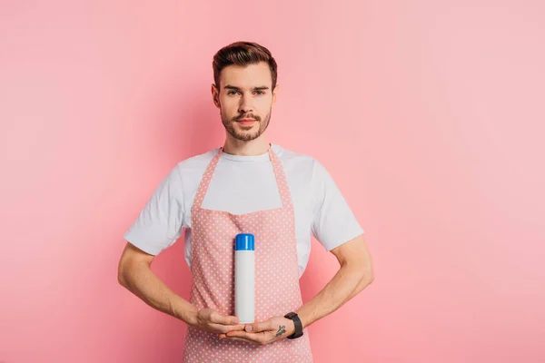 Serious young man in apron holding air freshener on pink background — Stock Photo