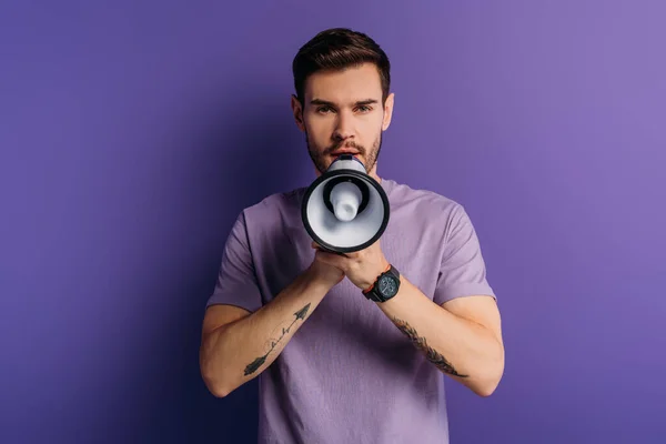 Handsome young man speaking in megaphone while looking at camera on purple background — Stock Photo