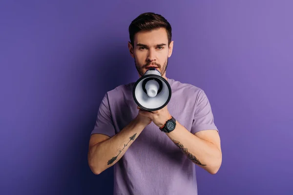 Schöner junger Mann spricht im Megafon, während er in die Kamera auf lila Hintergrund schaut — Stockfoto