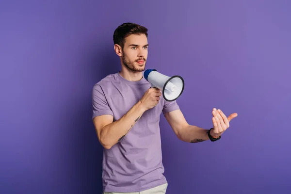 Серьезный молодой человек показывает прийти сюда жест, держа мегафон на фиолетовом фоне — стоковое фото
