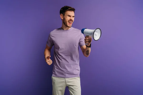 Роздратований молодий чоловік кричить в мегафоні, дивлячись на фіолетовий фон — Stock Photo