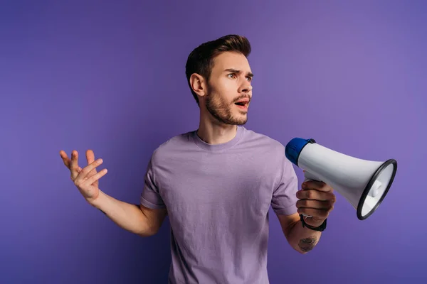 Sorprendido joven hombre sosteniendo megáfono y mirando hacia otro lado sobre fondo púrpura — Stock Photo