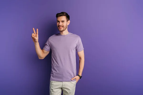 Sorridente jovem mostrando gesto de vitória enquanto segurando mão no bolso no fundo roxo — Fotografia de Stock