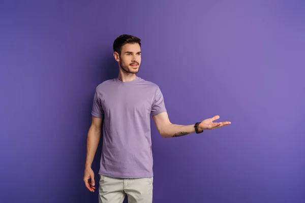 Bel homme positif debout avec le bras ouvert tout en regardant loin sur fond violet — Photo de stock