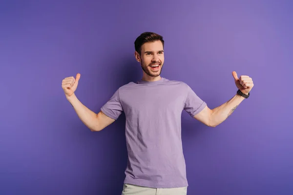 Возбужденный молодой человек показывает большие пальцы вверх, отводя взгляд на фиолетовом фоне — стоковое фото