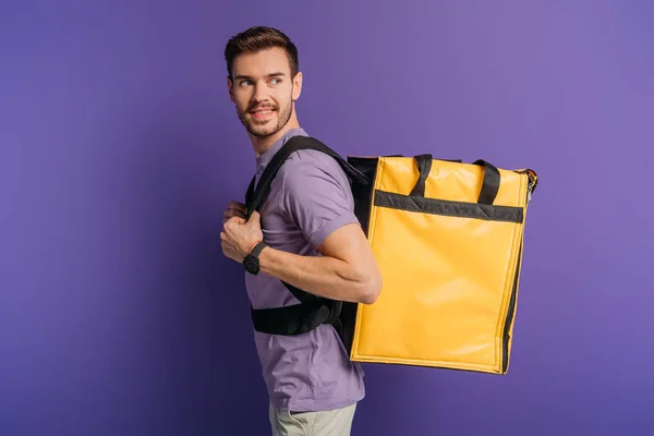 Cheerful delivery man looking away while carrying thermo backpack on purple background — Stock Photo