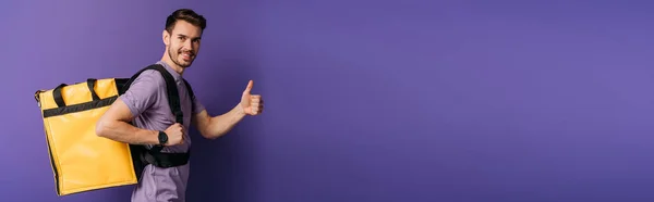 Panoramic shot of happy delivery man showing thumb up while looking at camera on purple background — Stock Photo