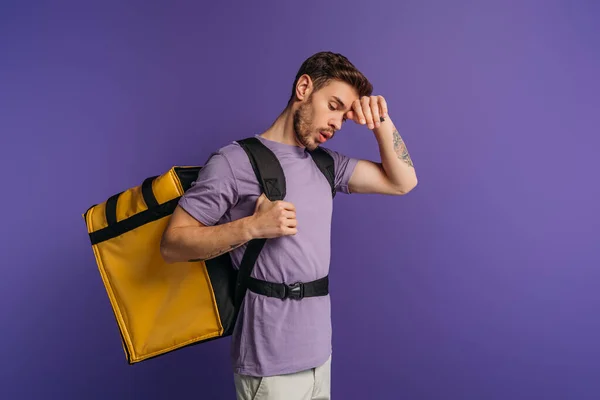 Exhausted delivery man touching forehead while carrying thermo backpack isolated on purple — Stock Photo