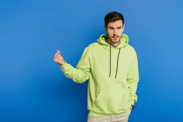 Junger selbstbewusster Mann zeigt, wie viel Geld Geste, während Hand in Tasche auf blauem Hintergrund — Stockfoto