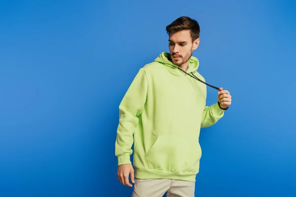 Handsome young man touching laces on hoodie while posing isolated on blue — Stock Photo
