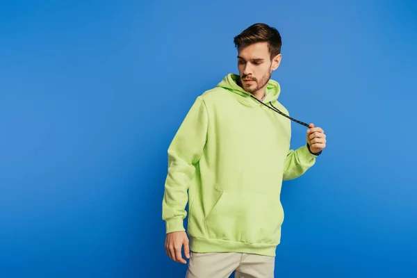 Guapo joven tocando cordones en sudadera con capucha mientras posando aislado en azul - foto de stock