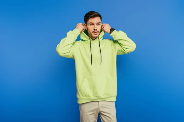 Handsome confident young man putting hood on while looking away on blue background — Stock Photo
