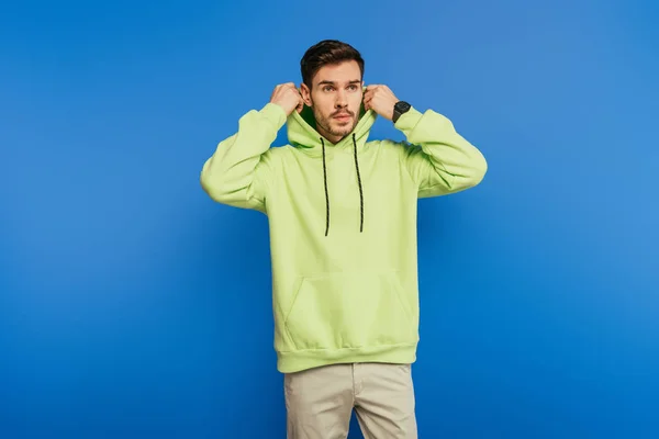 Guapo confiado joven hombre poniendo capucha mientras mira hacia otro lado en fondo azul - foto de stock