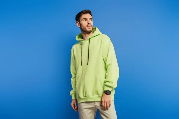 Guapo joven serio en sudadera con capucha mirando hacia otro lado sobre fondo azul - foto de stock