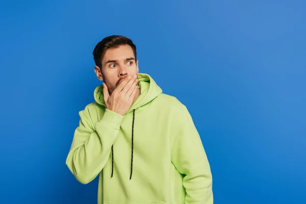 Schockierter junger Mann bedeckt Mund mit der Hand, während er isoliert auf blauem Grund wegschaut — Stockfoto