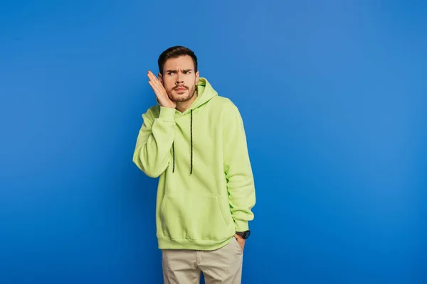 Curioso joven sosteniendo la mano cerca de la oreja mientras mira hacia otro lado aislado en azul - foto de stock