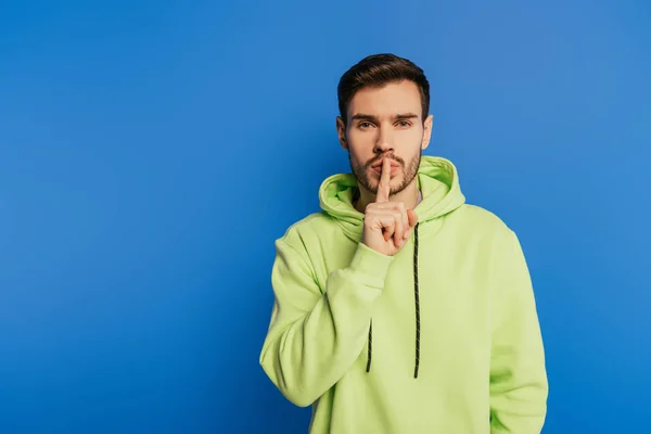 Serious young man showing hush gesture while looking at camera isolated on blue — Stock Photo