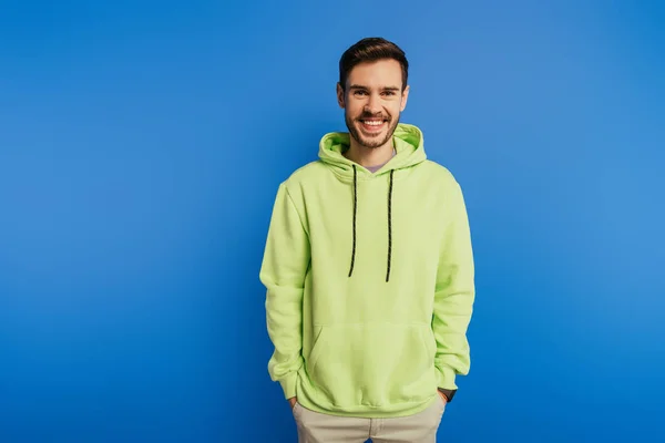Cheerful handsome man in hoodie smiling at camera while holding hands in pockets isolated on blue — Stock Photo