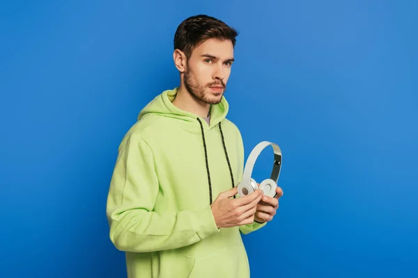 Pensativo joven sosteniendo auriculares inalámbricos mientras mira hacia otro lado sobre fondo azul - foto de stock