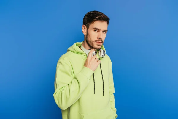 Serious young man touching wireless headphones while looking at camera isolated on blue — Stock Photo