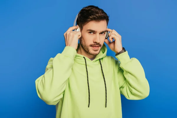 Confident young man touching wireless headphones while looking at camera isolated on blue — Stock Photo