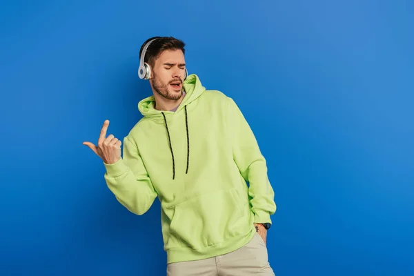 Jovem animado em fones de ouvido sem fio cantando com olhos fechados no fundo azul — Fotografia de Stock