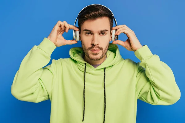 Serious young man touching wireless headphones while looking at camera isolated on blue — Stock Photo