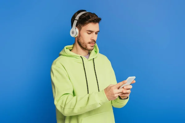 Jeune homme attentif dans les écouteurs sans fil en utilisant smartphone isolé sur bleu — Photo de stock