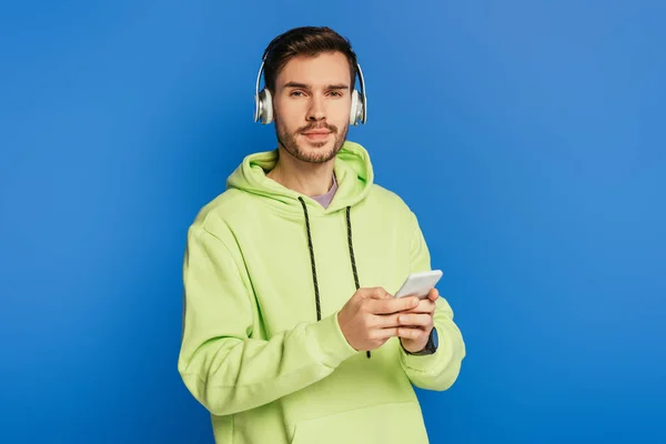 Thoughtful young man in wireless headphones holding smartphone and looking at camera isolated on blue — Stock Photo