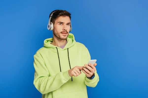 Thoughtful young man in wireless headphones using smartphone and looking away isolated on blue — Stock Photo