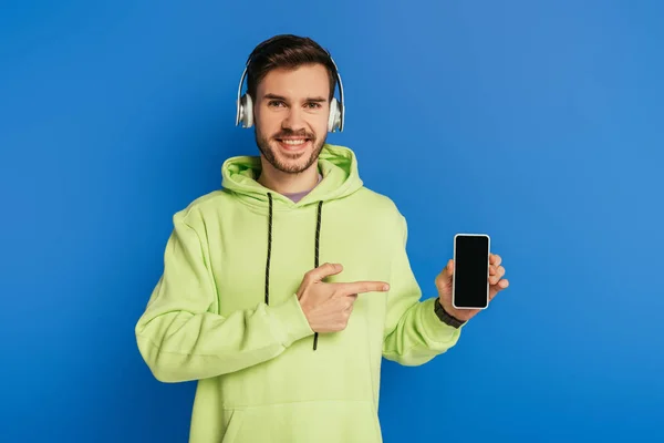 Joven feliz en auriculares inalámbricos apuntando con el dedo al teléfono inteligente con pantalla en blanco aislado en azul — Stock Photo