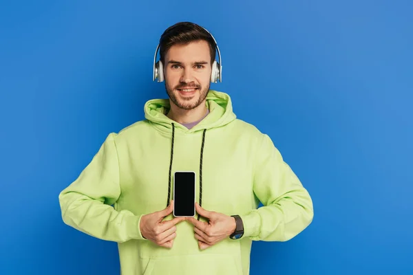 Allegro giovane uomo in cuffie wireless che mostra smartphone con schermo bianco isolato su blu — Foto stock