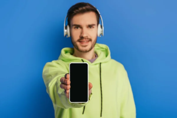 Selective focus of cheerful young man in wireless headphones showing smartphone with blank screen on blue background — Stock Photo