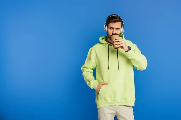 Verträumter junger Mann mit drahtlosen Kopfhörern, Kaffee to go trinkend und isoliert auf blauem Grund wegschauend — Stockfoto