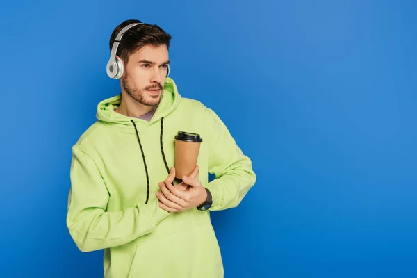 Verträumter junger Mann mit drahtlosen Kopfhörern, Kaffee to go in der Hand und isoliert auf blauem Grund schauend — Stockfoto
