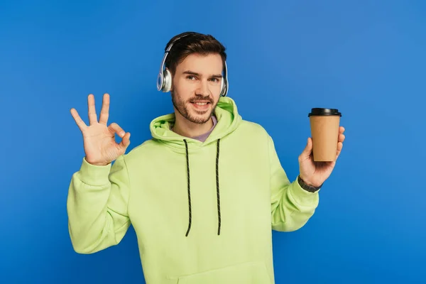 Happy young man in wireless headphones holding coffee to go and showing ok sign isolated on blue — Stock Photo