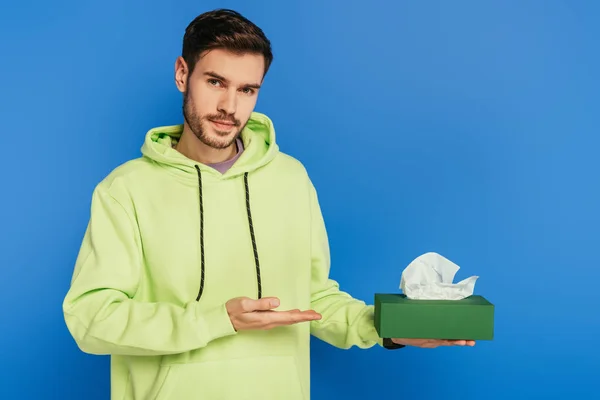 Schöner junger Mann zeigt mit der Hand auf Packung mit Papierservietten auf blauem Hintergrund — Stockfoto