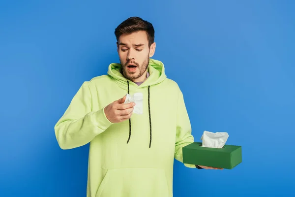 Sick man sneezing while holding paper napkins on blue background — Stock Photo