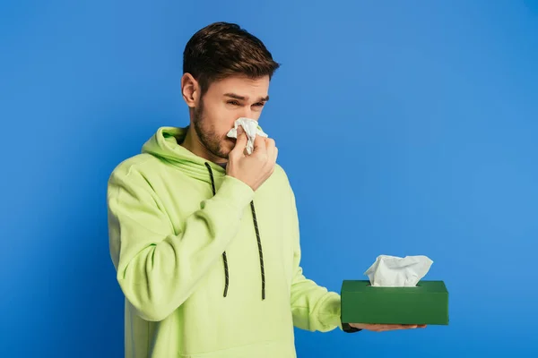 Jovem doente esfregando o nariz com guardanapo de papel no fundo azul — Fotografia de Stock