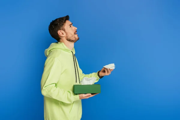 Vista lateral do homem triste olhando para cima e chorando enquanto segurando guardanapo de papel isolado no azul — Fotografia de Stock