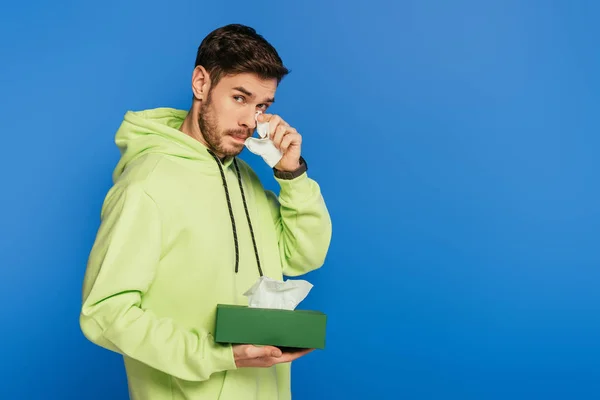 Chateado jovem chorando e enxugando lágrimas com guardanapo de papel isolado em azul — Fotografia de Stock