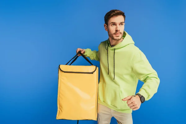 Serious delivery man looking away while holding thermo backpack isolated on blue — Stock Photo