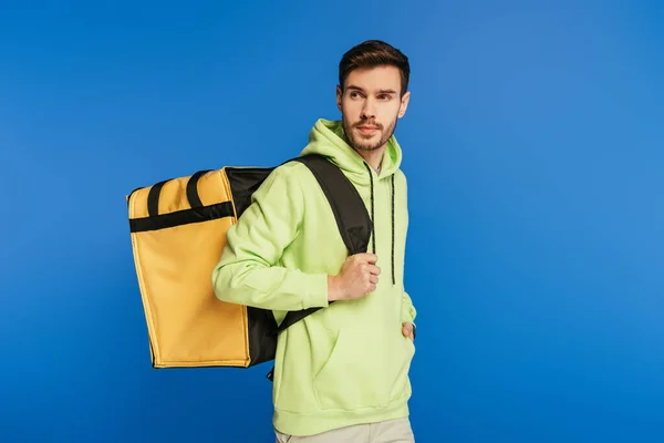 Schöner Lieferant trägt Thermo-Rucksack, während er isoliert auf blau wegschaut — Stockfoto