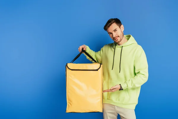 Guapo repartidor apuntando con la mano a la mochila termo sobre fondo azul - foto de stock