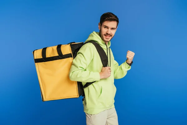 Happy delivery man showing winner gesture while carrying thermo backpack isolated on blue — Stock Photo